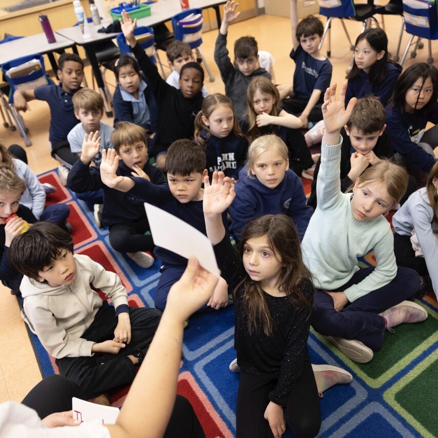 Elementary students seated on a colorful classroom rug, attentively raising their hands to answer a question from their teacher during a group learning activity at Penn Alexander School.